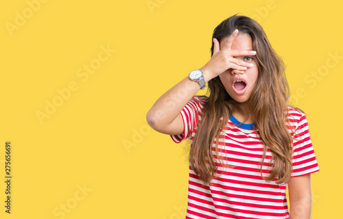 Young beautiful brunette woman wearing stripes t-shirt over isolated background peeking in shock covering face and eyes with hand, looking through fingers with embarrassed expression.