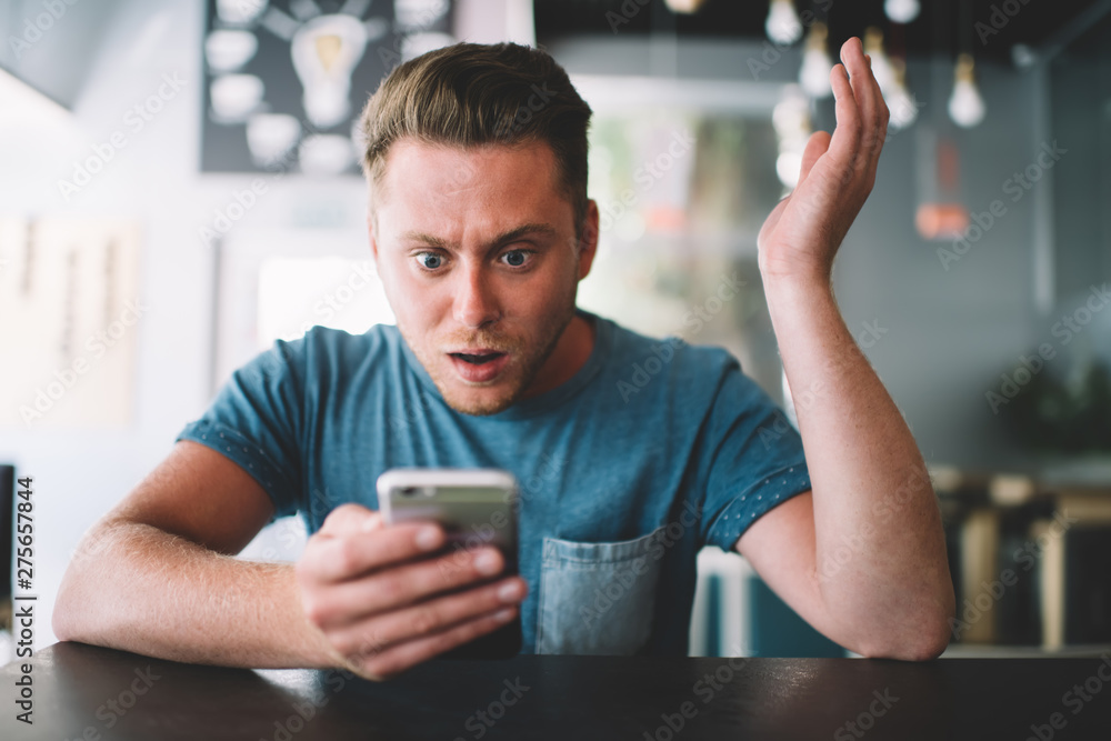 Amazed hipster guy with cellphone gadget in hands feeling shocked from notification on application sitting at cafe interior with wifi internet, wonder Caucasian male blogger reading text comment