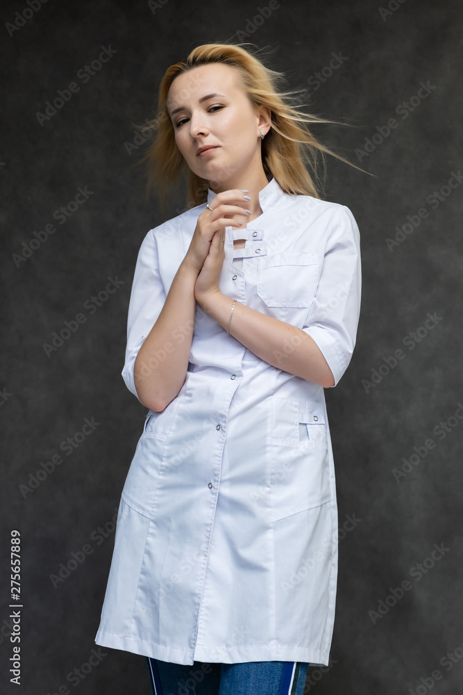 Portrait of a knee-high pretty interesting beautiful girl blonde doctor on a gray background in a white medical coat. Standing in front of the camera, smiling, a lot of emotions. Portrait of a knee-high pretty interesting beautiful girl blonde doctor on a gray background in a white medical coat. Standing in front of the camera, smiling, a lot of emotions.