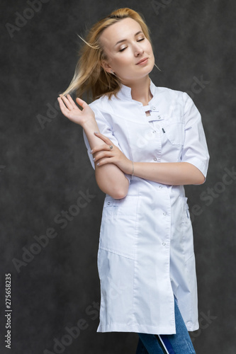 Portrait of a knee-high pretty interesting beautiful girl blonde doctor on a gray background in a white medical coat. Standing in front of the camera, smiling, a lot of emotions. © Вячеслав Чичаев