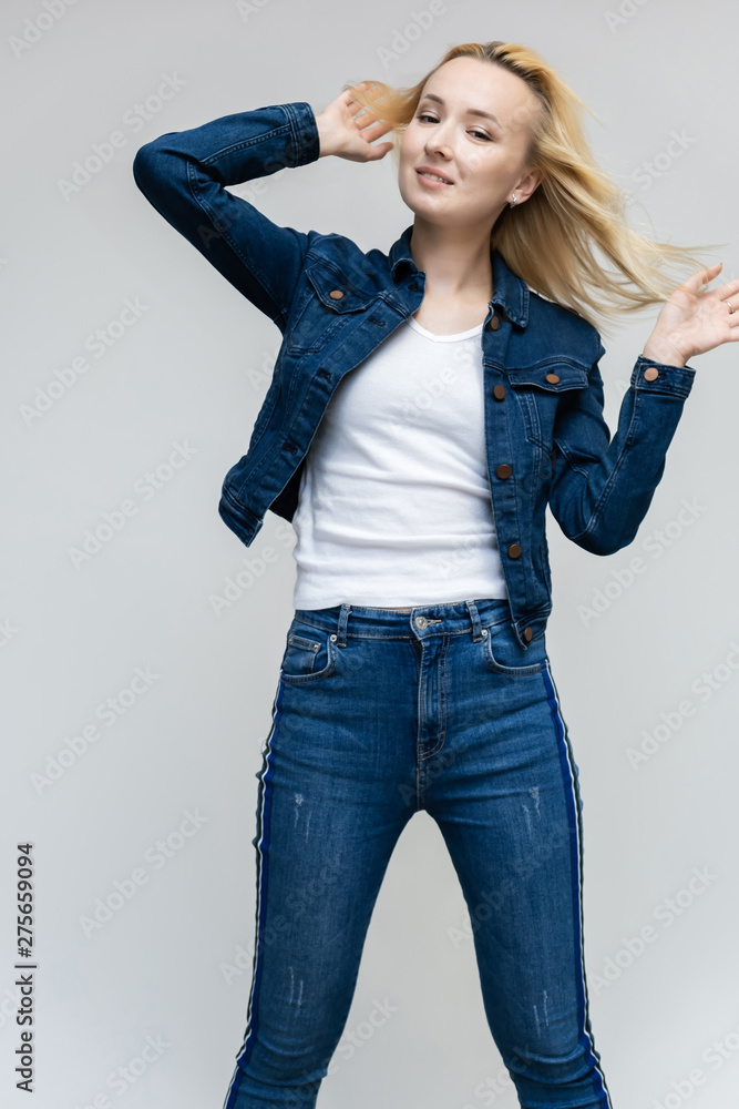 Portrait of pretty interesting fashionable blonde girl on a white background in jeans and a blue jacket. Standing in front of the camera, smiling, showing hands. Shows a lot of emotions. Portrait of pretty interesting fashionable blonde girl on a white background in jeans and a blue jacket. Standing in front of the camera, smiling, showing hands. Shows a lot of emotions.
