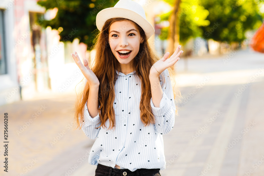 Emotional girl teenager in a hat walks through the summer city. Debonair caucasian girl enjoying photoshoot on city background. Emotional girl teenager in a hat walks through the summer city. Debonair caucasian girl enjoying photoshoot on city background.