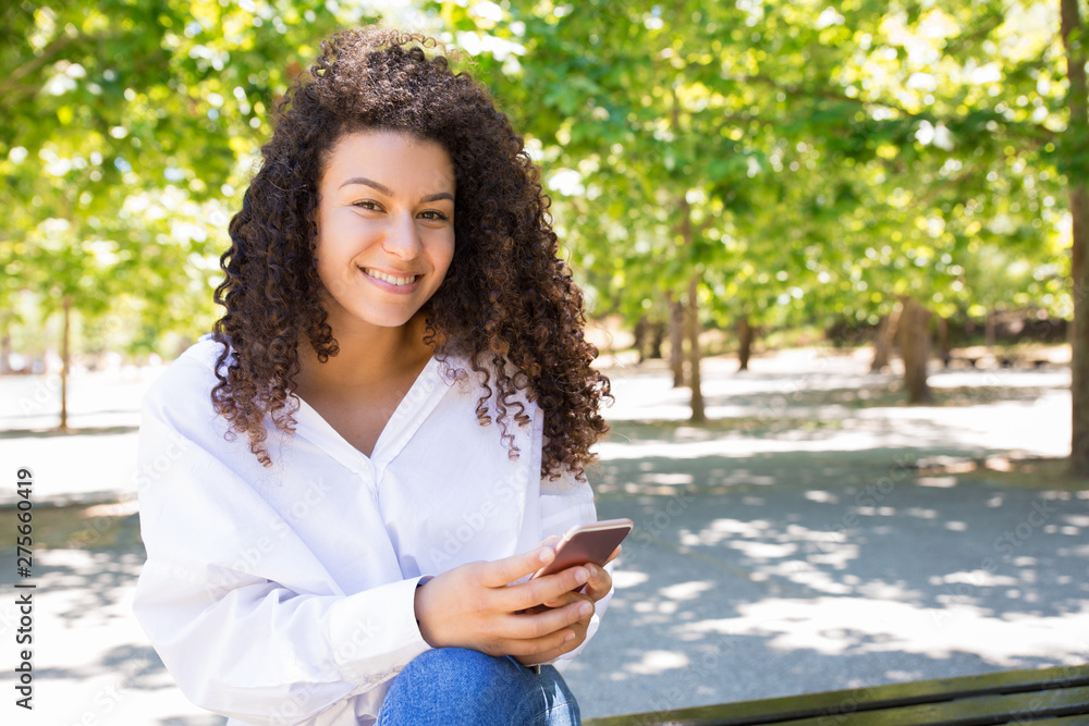 Happy pretty young woman using smartphone on bench in park. Beautiful lady wearing blouse and sitting with green trees in background. Communication and nature concept. Front view. Happy pretty young woman using smartphone on bench in park. Beautiful lady wearing blouse and sitting with green trees in background. Communication and nature concept. Front view.
