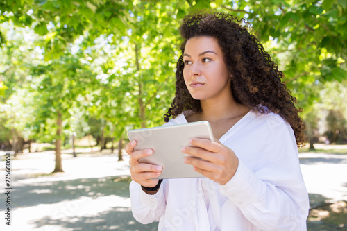 Pensive pretty young woman browsing on tablet in park. Beautiful lady wearing blouse and looking away with green trees in background. Technology and nature concept. Front view.