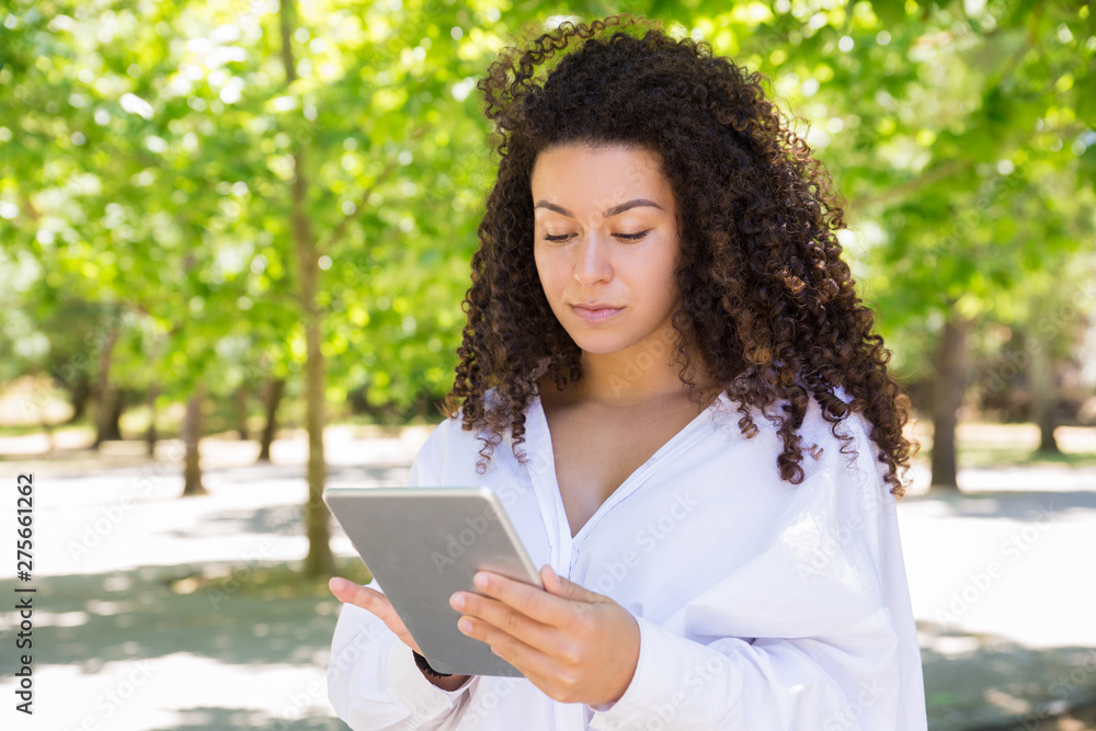 Serious pretty young woman browsing on tablet in park. Beautiful lady wearing blouse and standing with green trees in background. Technology and nature concept. Front view. Serious pretty young woman browsing on tablet in park. Beautiful lady wearing blouse and standing with green trees in background. Technology and nature concept. Front view.