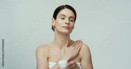 Portrait of the young Caucasian pretty woman wrapped in the white towel moisturizing her neck with a cream and holding a cream on hand while demonstating it to camera. photo