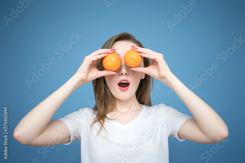 Young beautiful girl model with oranges on blue background, holding oranges between eyes, mouth open in surprise and delight © Анастасия Семашко
