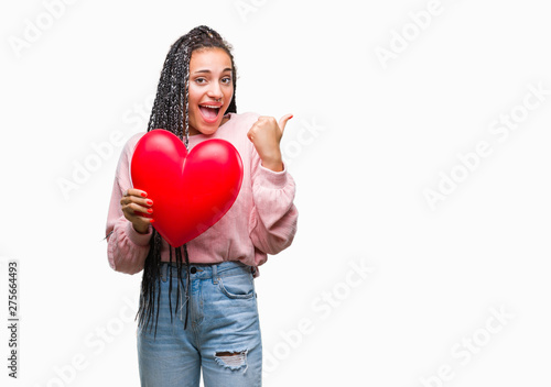 Young braided hair african american girl holding read heart over isolated background pointing and showing with thumb up to the side with happy face smiling © Krakenimages.com