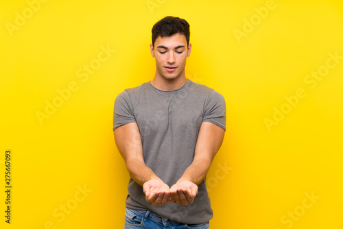 Handsome man over isolated yellow wall holding copyspace imaginary on the palm to insert an ad