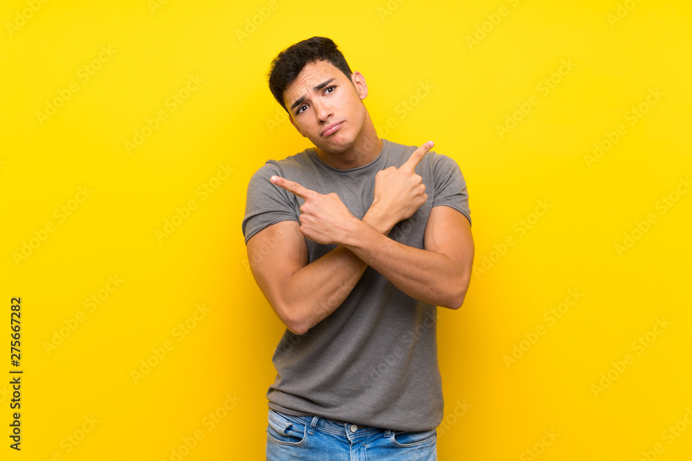Handsome man over isolated yellow wall pointing to the laterals having doubts Handsome man over isolated yellow wall pointing to the laterals having doubts