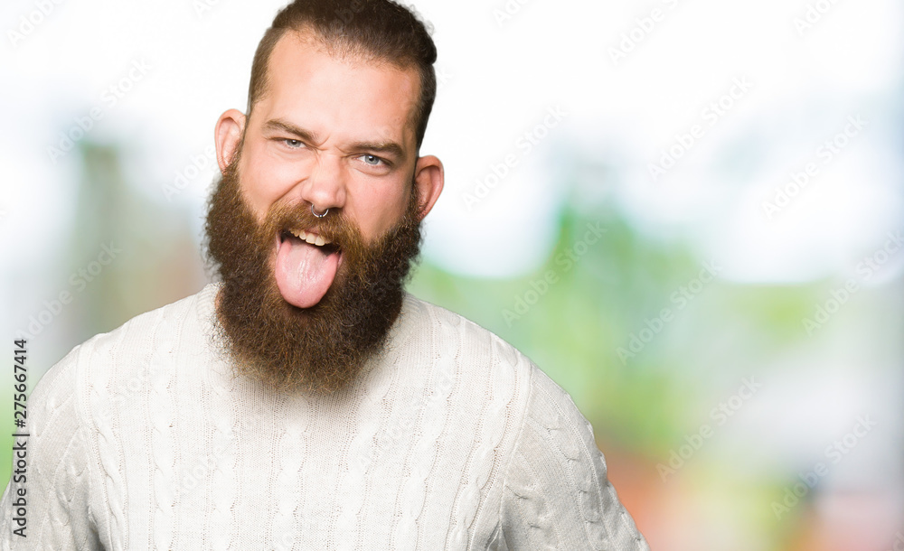 Fototapeta premium Young hipster man wearing winter sweater sticking tongue out happy with funny expression. Emotion concept.
