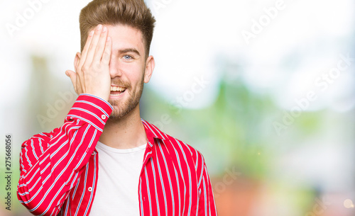Young handsome man wearing red shirt covering one eye with hand with confident smile on face and surprise emotion.