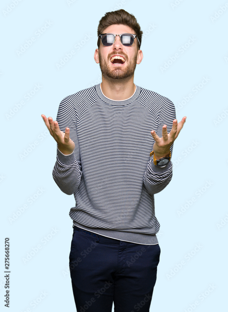 Naklejka premium Young handsome man wearing sunglasses crazy and mad shouting and yelling with aggressive expression and arms raised. Frustration concept.