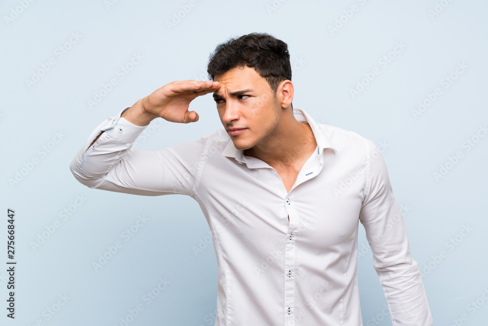 Handsome man over blue wall looking far away with hand to look something Handsome man over blue wall looking far away with hand to look something