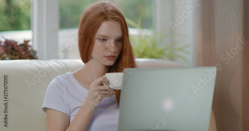 Attractive girl sitting at home on the couch using modern laptop and drinking coffee. tracking shot. in slow motion. Shot on Canon 1DX mark2 4K camera photo