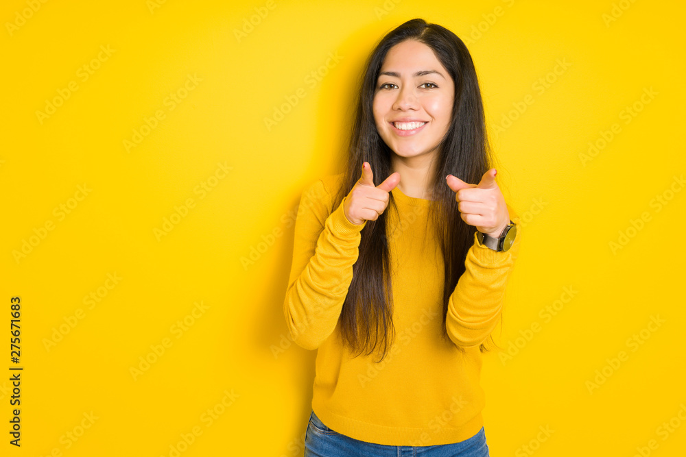 Beautiful brunette woman over yellow isolated background pointing fingers to camera with happy and funny face. Good energy and vibes. Beautiful brunette woman over yellow isolated background pointing fingers to camera with happy and funny face. Good energy and vibes.