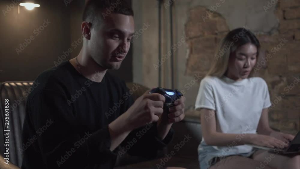 Handsome young guy having fun on evening emotionally playing video games near his girlfriend working on laptop in modern loft club Handsome young guy having fun on evening emotionally playing video games near his girlfriend working on laptop in modern loft club