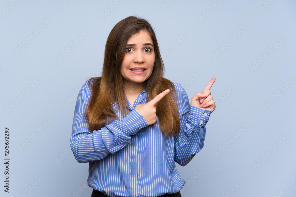 Young girl over isolated blue wall frightened and pointing to the side Young girl over isolated blue wall frightened and pointing to the side