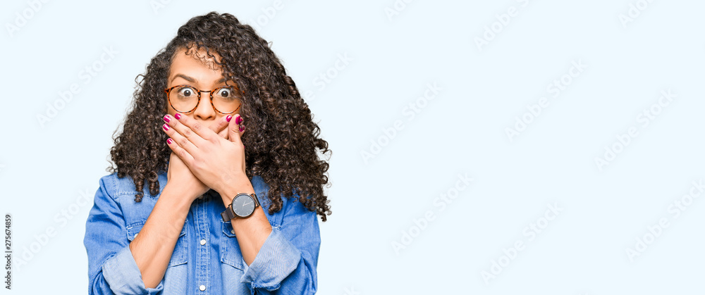 Young beautiful woman with curly hair wearing glasses shocked covering mouth with hands for mistake. Secret concept. Young beautiful woman with curly hair wearing glasses shocked covering mouth with hands for mistake. Secret concept.