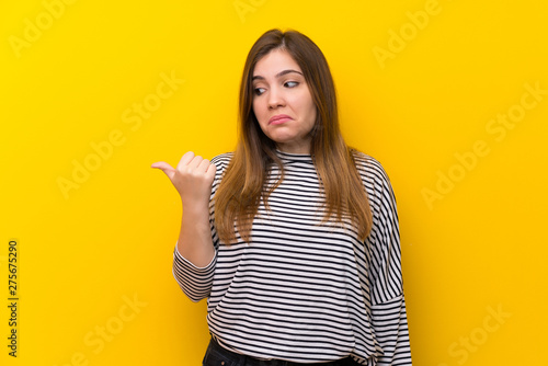 Young girl over yellow wall unhappy and pointing to the side