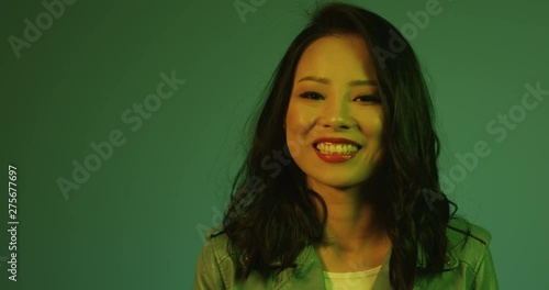 Portrait of the beautiful Asian young woman looking straight to the camera and smiling joyfully on the wall background with green light. Close up. photo