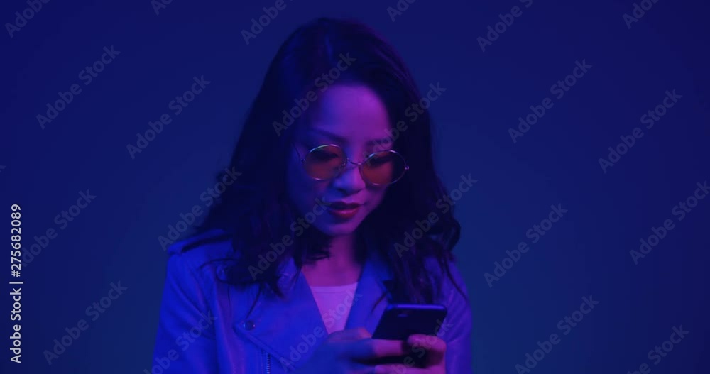Portrait shot of the charming and beautiful young Asian woman in sunglasses tapping and texting on the smartphone and then smiling to the camera on the dark blue neon light. Close up. Portrait shot of the charming and beautiful young Asian woman in sunglasses tapping and texting on the smartphone and then smiling to the camera on the dark blue neon light. Close up.