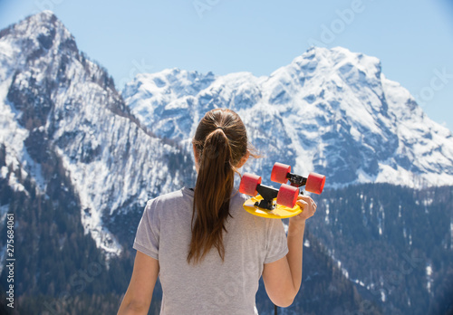 A young woman with blonde hair standing on a background of mountains and holding a skateboard photo