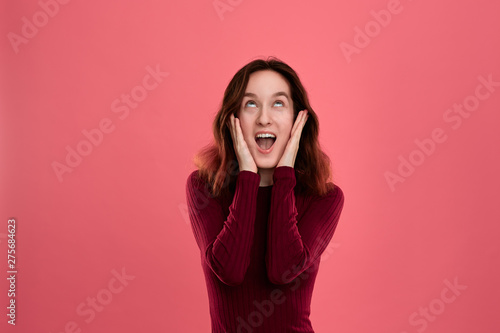 Surprised young lady standing isolated over dark pink background with hands raised close to mouth expressing emotion of shock.