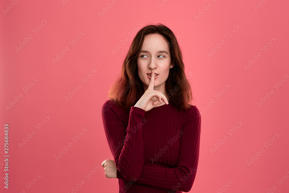 Beautiful young lady standing isolated over dark pink background with finger raised close to mouth expressing gesture of keeping silence and looking at the camera. Beautiful young lady standing isolated over dark pink background with finger raised close to mouth expressing gesture of keeping silence and looking at the camera.