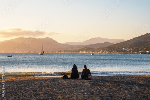 Seascape with a young couple on the sandy beach of a bay with the coastline on the horizon at sunset, Sestri Levante, Genoa, Liguria, Italy