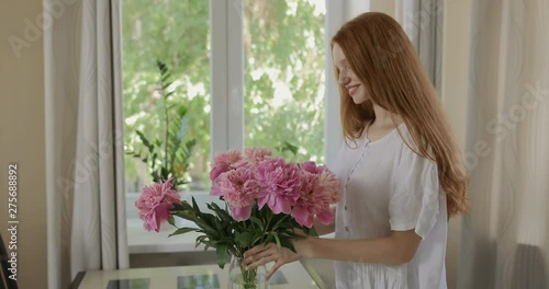 Attractive girl put the flowers of peonies in a vase and corrects the bouquet. Red-haired woman enjoying the fresh flowers and aroma of peonies. tracking shot. in slow motion. Shot on Canon 1DX mark2  photo