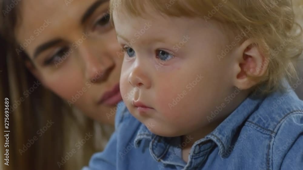 Tilting close-up shot of doting Caucasian mother posing for camera with her sulky blond baby boy, talking to him gently and encouraging him to smile for camera without success Tilting close-up shot of doting Caucasian mother posing for camera with her sulky blond baby boy, talking to him gently and encouraging him to smile for camera without success
