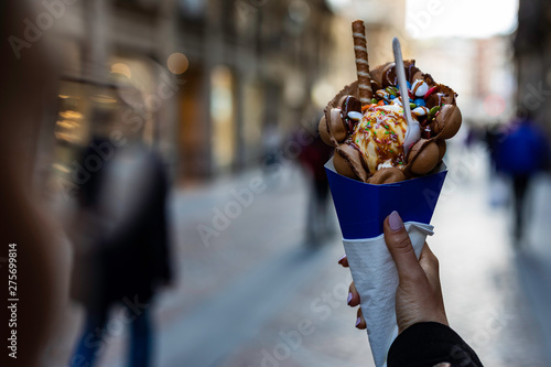 Woman hand holding a bubble waffle with ice cream and candies on a blue paper cone with blurred unrecognizable crowd on the background photo