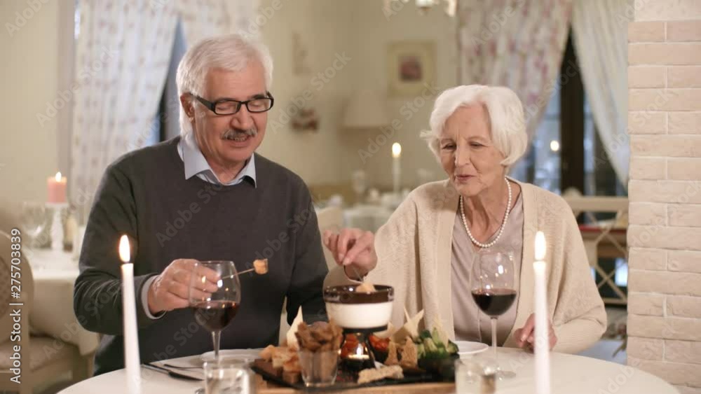Waist-up shot of retired 60-something Caucasian husband and wife enjoying dinner in candlelit restaurant, carefully dipping fondue snacks into melted cheese and tasting them Waist-up shot of retired 60-something Caucasian husband and wife enjoying dinner in candlelit restaurant, carefully dipping fondue snacks into melted cheese and tasting them