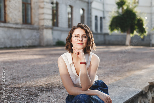 Beautiful young brunette woman with short wavy hair, wearing white sleeveless shirt, blue skirt and glasses, sitting outdoors in sunny day. © NesolenayaAleksandra