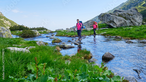 Two active, fit women hikers crossing a mountain river by stepping on rocks, with heavy camping backpacks and trekking poles - Bordul Tomii in Retezat mountains (Carpathians), Romania.