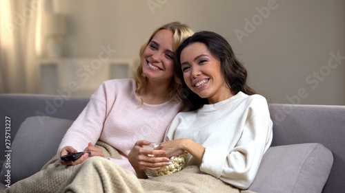 Smiling female friends with popcorn watching comedy show at home, evening