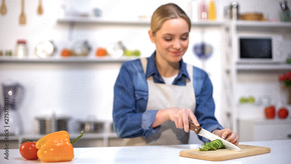 Beautiful young lady slicing cucumber, preparing salad, vegetarian lifestyle