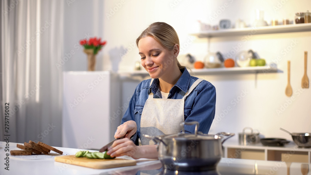 Smiling woman slicing cucumber in kitchen, preparing healthy low-caloried salad