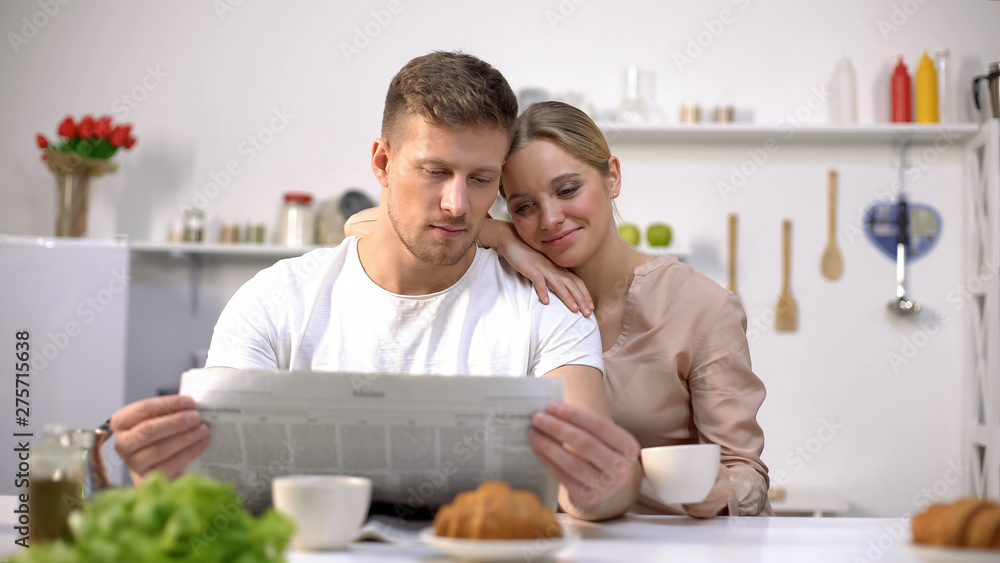 Young husband reading newspaper with wife holding coffee cup, morning together