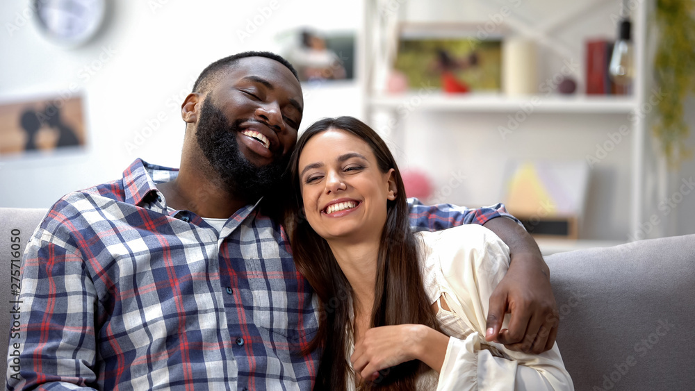Fototapeta premium Nice multiethnic couple smiling and sitting on sofa at home, having fun together