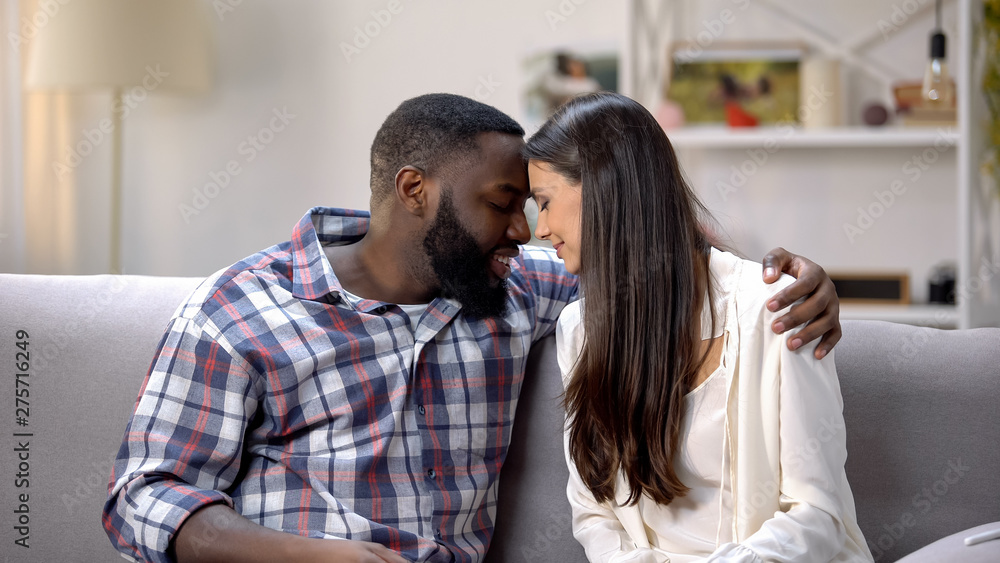 Loving mixed-race couple touching foreheads and hugging on couch, tenderness Loving mixed-race couple touching foreheads and hugging on couch, tenderness