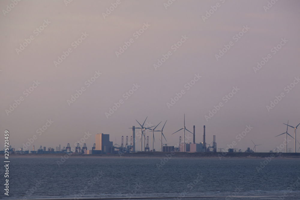 Energy import. Industrial and harbor area in the evening light from a distance. In between wind turbines and water in the foreground.