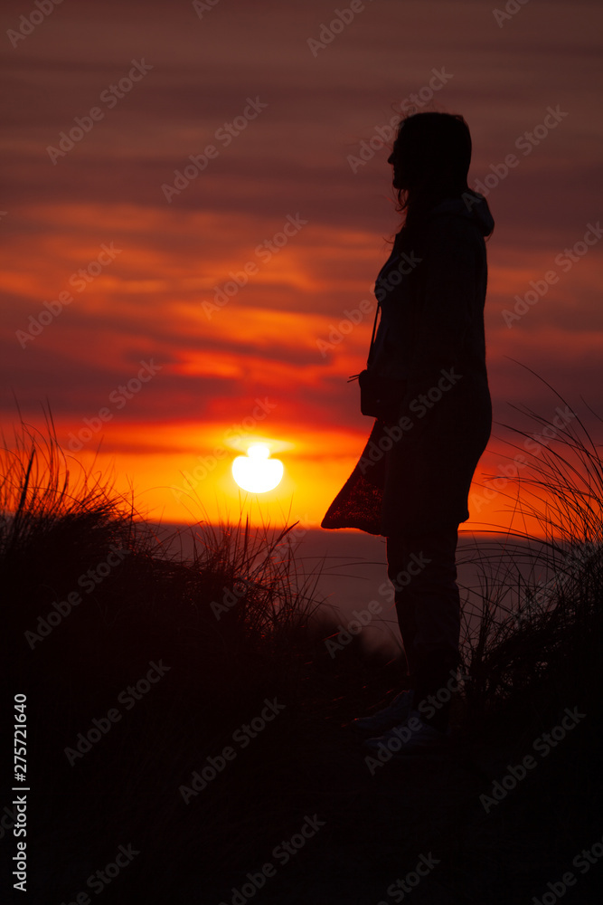 Woman on a dune in the sunset.Silhouette of person and dune grass with low sun and glowing red sky