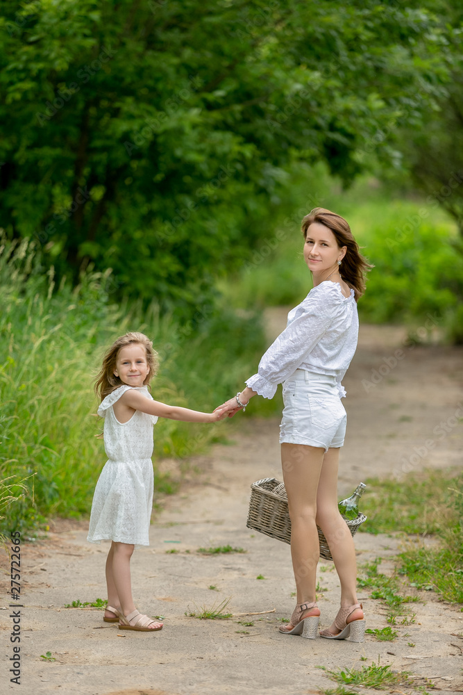 Fototapeta premium Beautiful young mother and her little daughter in white dress having fun in a picnic. They stand on a road in the park, holding hands, and look into the camera. Maternal care and love. Vertical photo