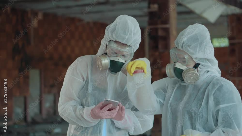 Two girls scientists in a white protective suit and respirator enter the data on the level of radiation into the phone. The hazardous substance in a closed test tube is checked in an abandoned room. 4 Two girls scientists in a white protective suit and respirator enter the data on the level of radiation into the phone. The hazardous substance in a closed test tube is checked in an abandoned room. 4
