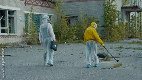 Checking the level of radiation in an abandoned territory. A scientist in a yellow protective suit and respirator holds a dosimeter. Two students in white defense walk with a suitcase, , collect evide photo