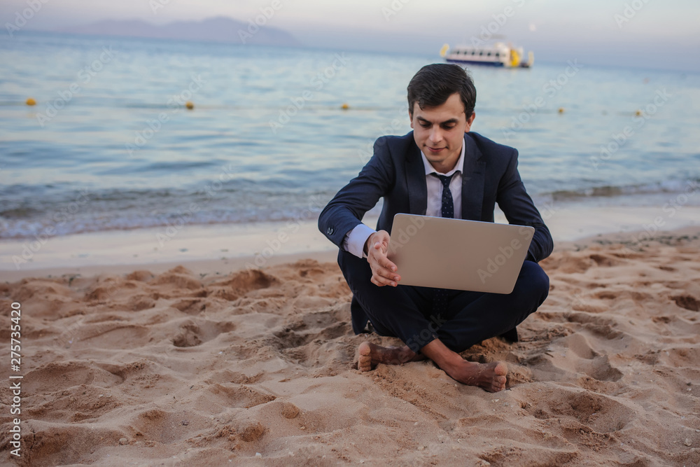 close up photo of a young man in suit with laptop working on the beach and talking to someone