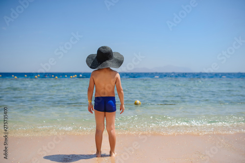 back view photo of a 7-year boy in blue underwear and a blue hat on the beach looking at the sea