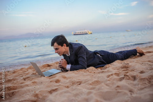 close up photo of a young man in suit with laptop lies on the beach and talking to someone on computer photo